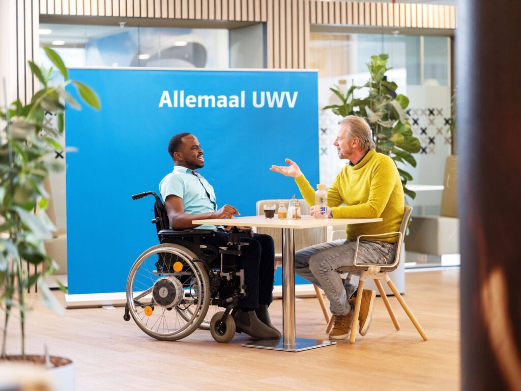 Yuriandy en Martijn zitten samen achter een bureau. Ze lachen naar elkaar. Yuriandy is een Zwarte man met een lichtblauw overhemd. Martijn is een witte man en draagt een gele trui. Op de achtergrond staat een groot blauw bord met daarop de tekst 'Allemaal UWV'.