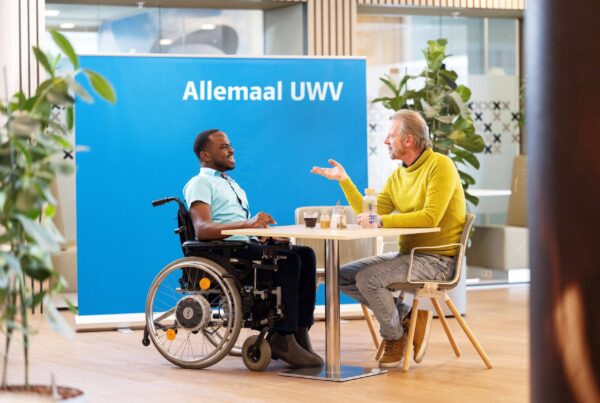 Yuriandy en Martijn zitten samen achter een bureau. Ze lachen naar elkaar. Yuriandy is een Zwarte man met een lichtblauw overhemd. Martijn is een witte man en draagt een gele trui. Op de achtergrond staat een groot blauw bord met daarop de tekst 'Allemaal UWV'.