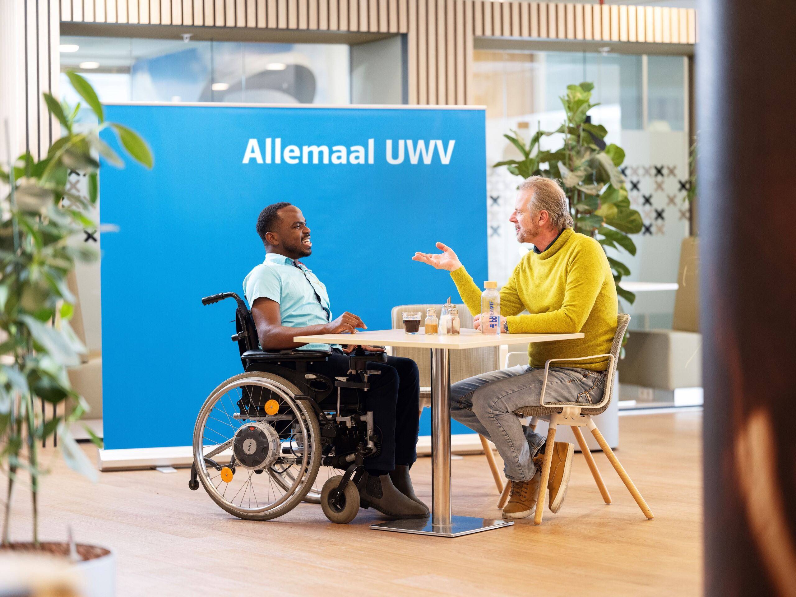 Yuriandy en Martijn zitten samen achter een bureau. Ze lachen naar elkaar. Yuriandy is een Zwarte man met een lichtblauw overhemd. Martijn is een witte man en draagt een gele trui. Op de achtergrond staat een groot blauw bord met daarop de tekst 'Allemaal UWV'.